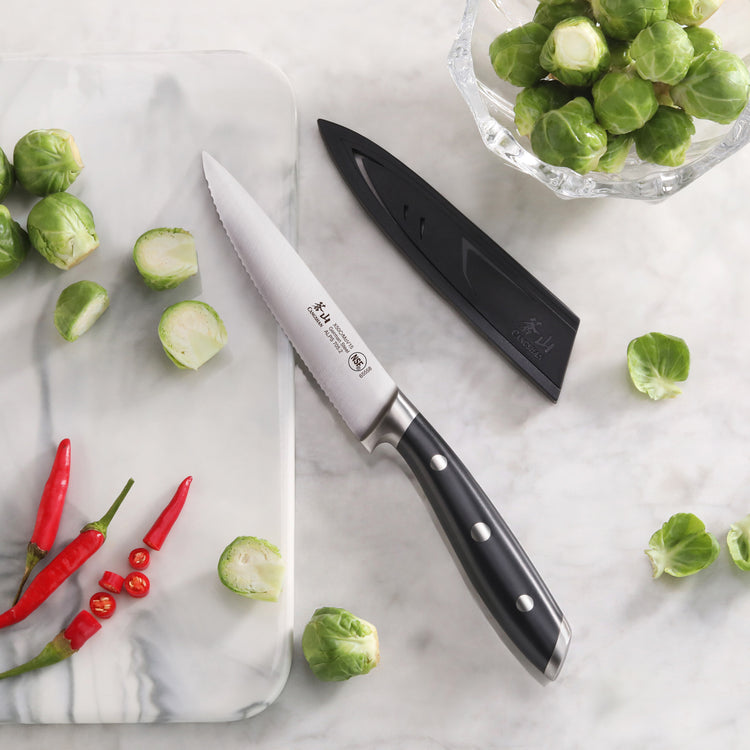 5-inch serrated utility knife with black handle and sheath next to sliced Brussels sprouts and red chili peppers on marble su