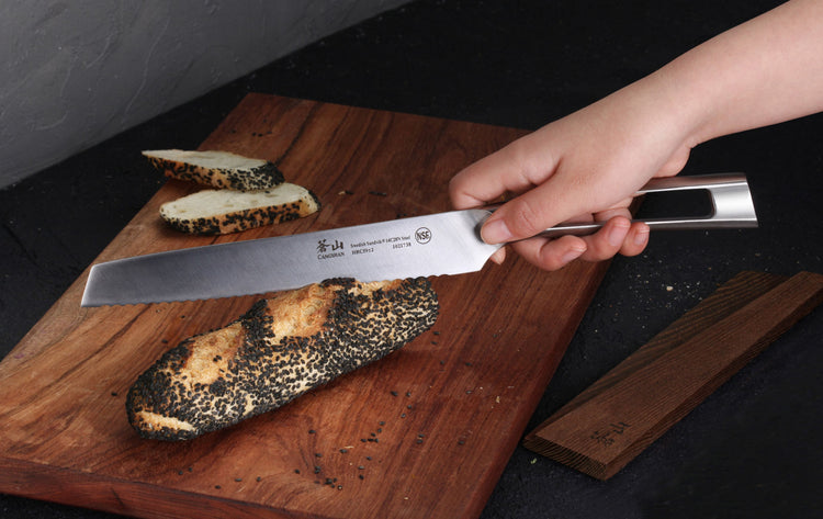 Hand holding a stainless steel bread knife slicing a burnt loaf on a wooden cutting board with an ash wood sheath nearby.