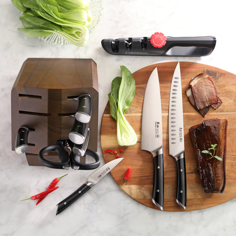 Knife block with six knives, kitchen scissors, knife sharpener, two pieces of cooked meat, bok choy, and red chili peppers on