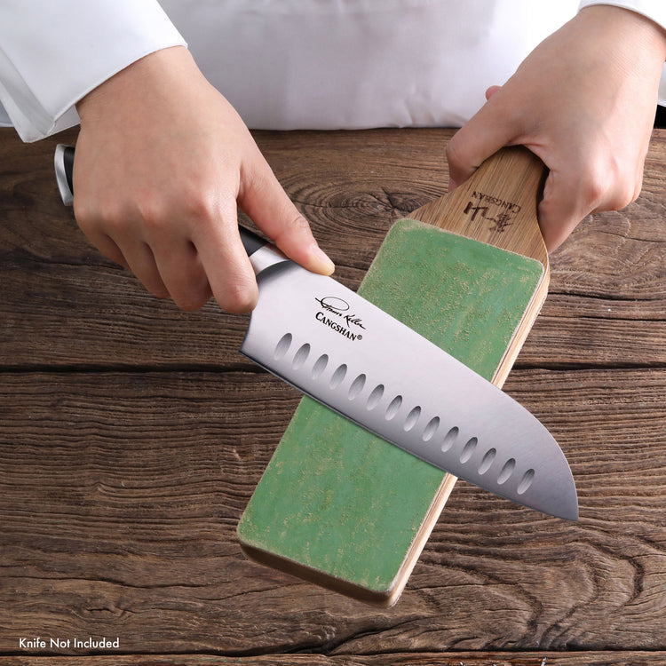Hands holding a sharpening stone and a knife blade being sharpened on a wooden surface.