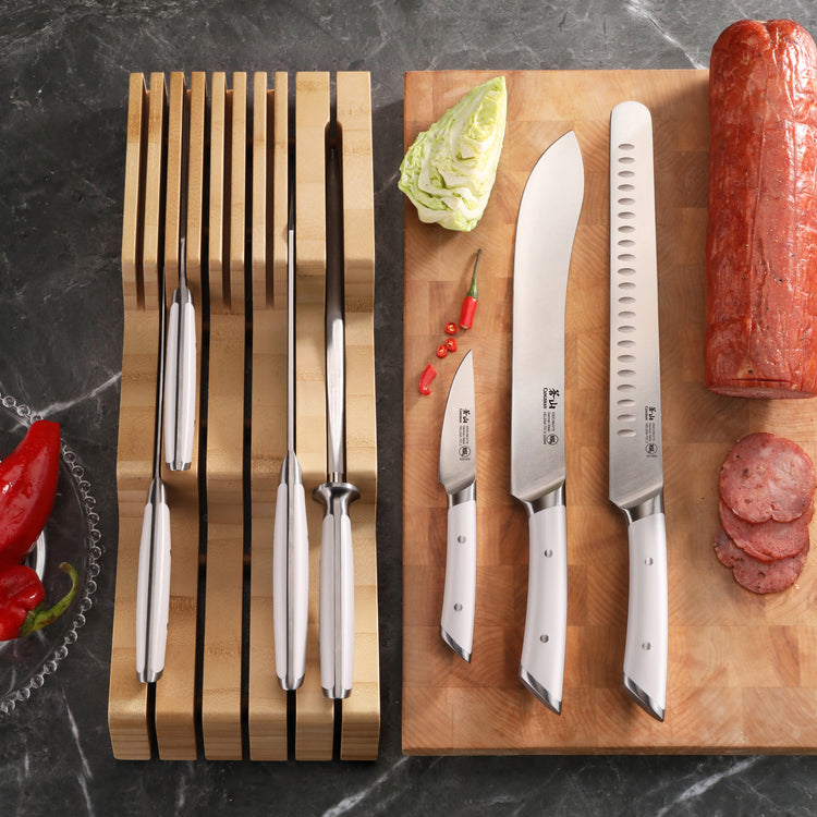 Set of eight stainless steel knives with white handles in a bamboo in-drawer organizer next to three knives on a wooden board