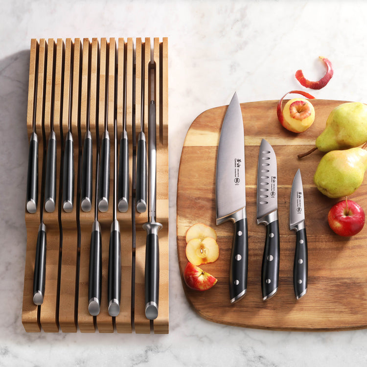 Fifteen black-handled knives in a bamboo in-drawer tray next to three knives on a wooden cutting board with sliced apples and