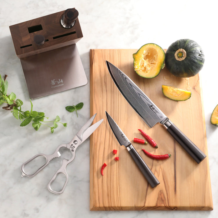 Two Damascus steel knives with dark handles on a wooden cutting board next to a halved green squash and red chili peppers, wi