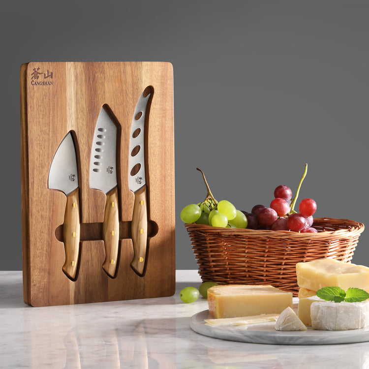 Three olive wood-handled cheese knives in an acacia wood board next to a basket of grapes and assorted cheeses.