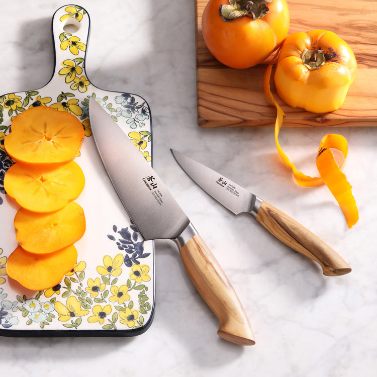 Two knives with wooden handles on a marble surface, one slicing a persimmon on a floral cutting board, the other next to peel