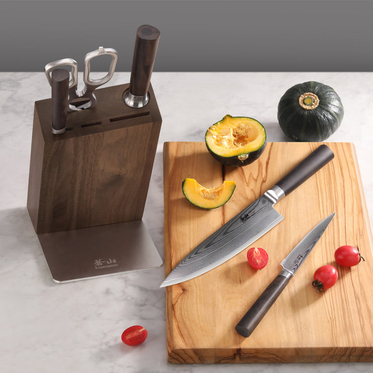 Knife block with three knives, two knives on a wooden cutting board, sliced squash, and cherry tomatoes on a marble surface.