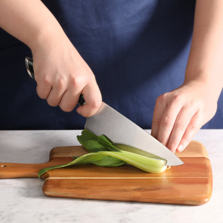 Hands using an 8-inch chef's knife to cut a bok choy on a wooden cutting board.