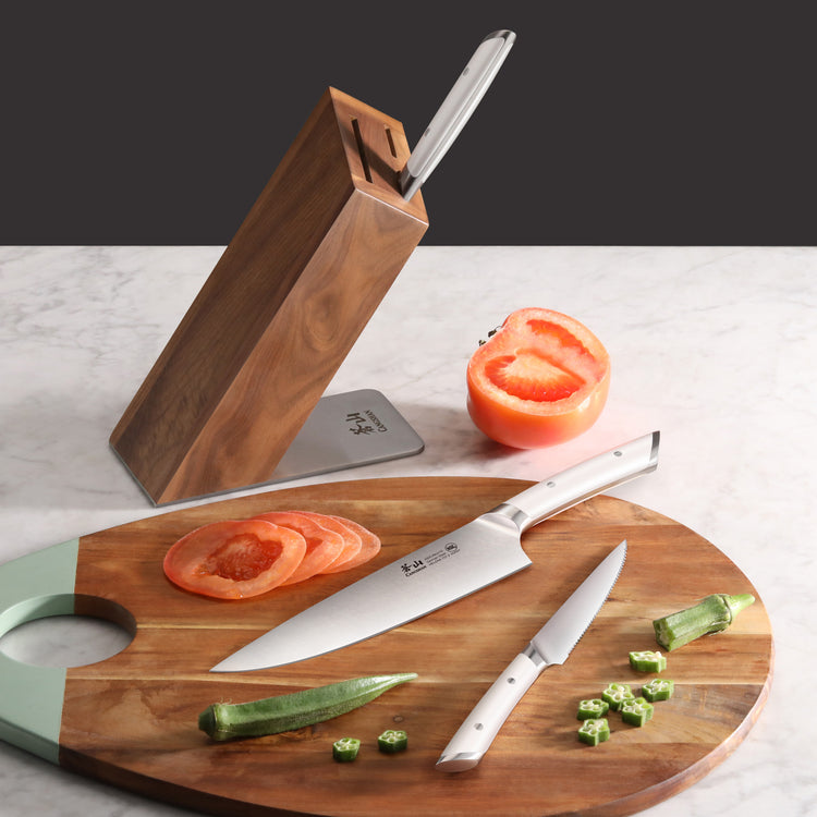 Wooden knife block with two stainless steel knives on a cutting board slicing tomato and okra.