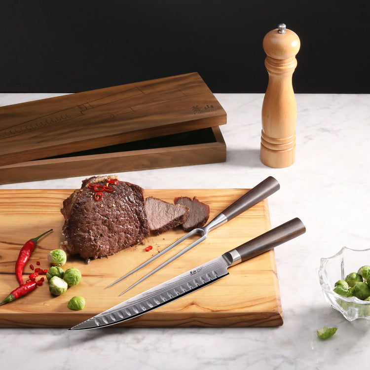 Carving knife and fork with dark handles on wooden board with sliced roast, chili peppers, Brussels sprouts, and walnut box.