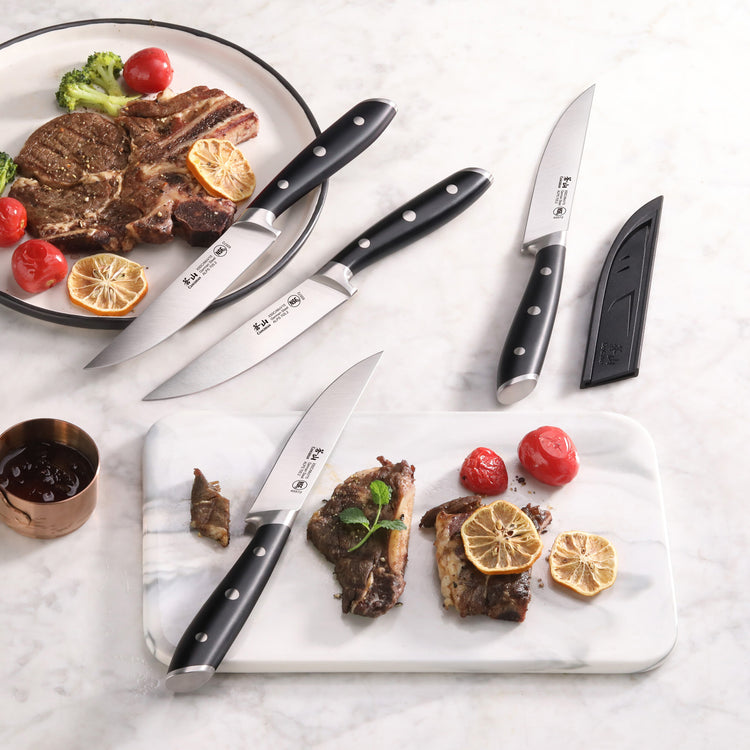 Four black-handled steak knives with stainless steel blades, two plates with cooked steak, grilled lemon, and cherry tomatoes