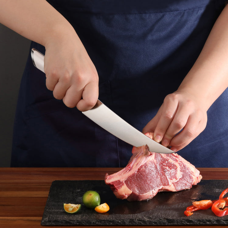Person using a boning knife to cut a raw piece of meat on a black cutting board with lime and sliced red peppers nearby.