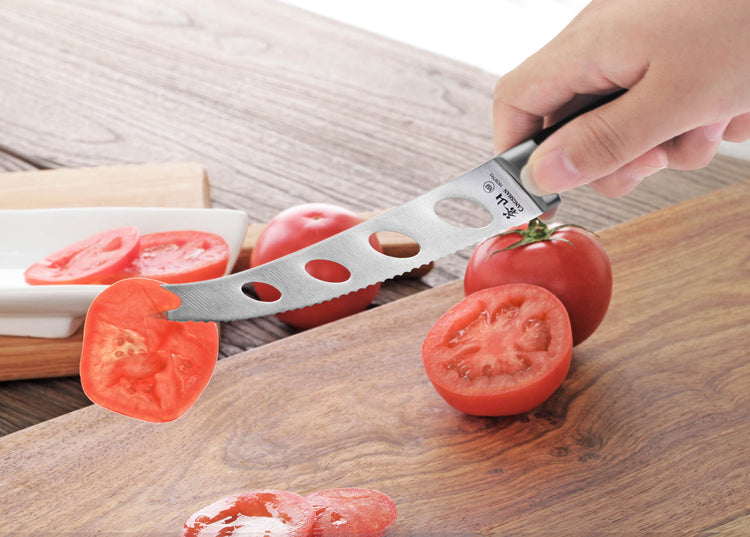 Hand holding a 5-inch tomato/cheese knife with holes, slicing a tomato on a wooden cutting board.