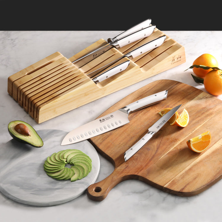 Seven-piece knife set with white handles in a bamboo in-drawer tray, displayed with sliced avocado and orange on cutting boar