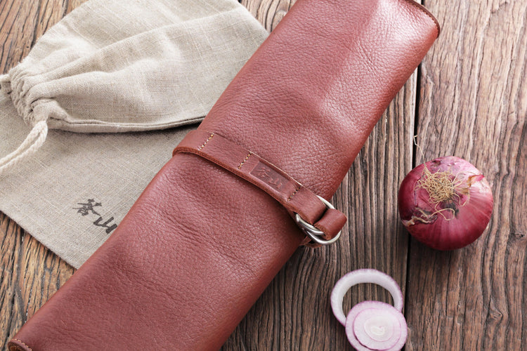 Brown leather roll knife case secured with a strap, placed on a wooden surface next to a beige drawstring pouch and a sliced