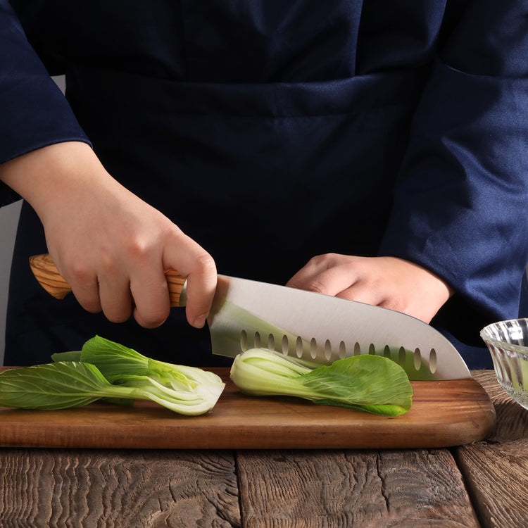 Person slicing bok choy on wooden board with a 7-inch Santoku knife featuring a wooden handle and scalloped blade.