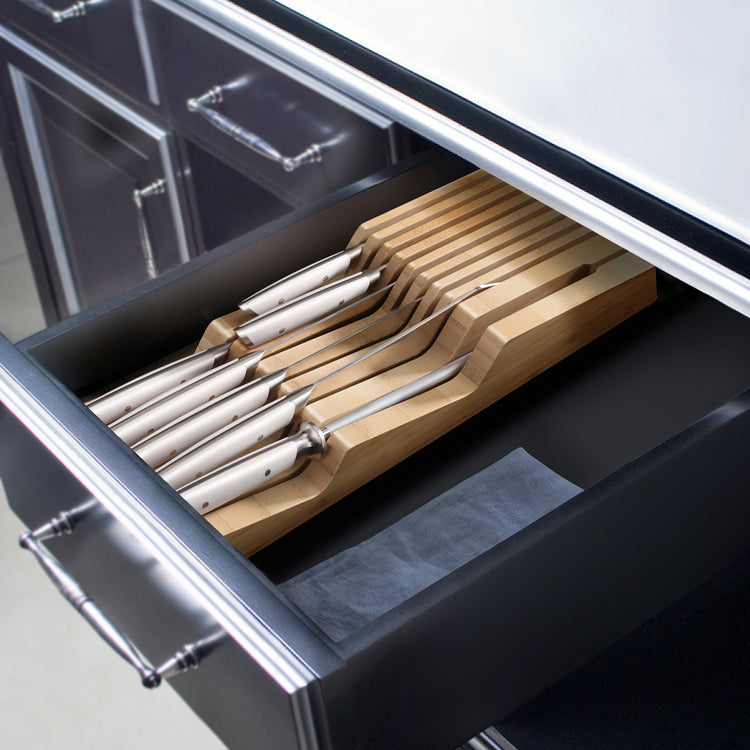 Eight stainless steel knives with white handles neatly arranged in a bamboo tray inside a kitchen drawer.