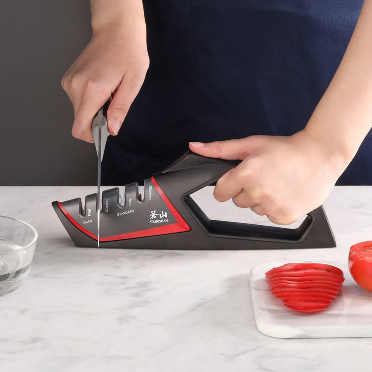 Hand holding a knife sharpening on a black and red handheld sharpener with coarse and fine slots on a marble surface.
