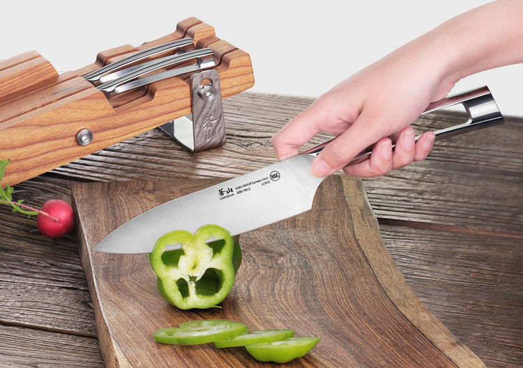 Hand holding a chef's knife slicing a green bell pepper on a wooden cutting board with a knife block in the background.