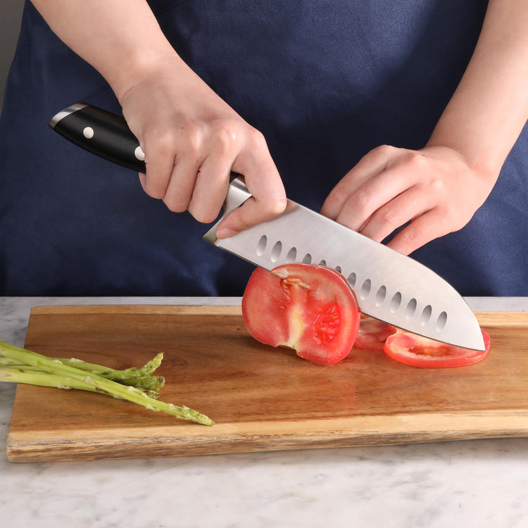 Hands slicing a tomato on a wooden cutting board with a black-handled Santoku knife, asparagus nearby.