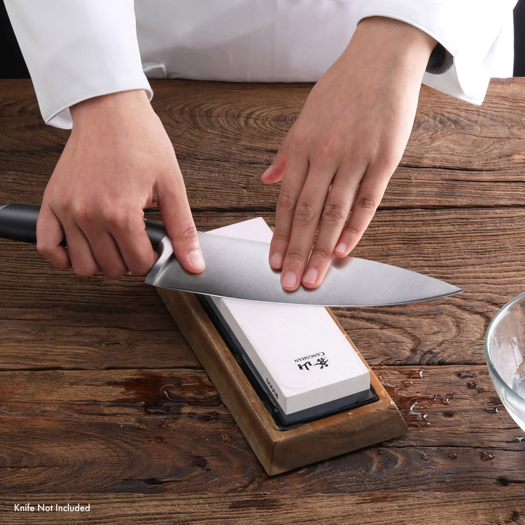 Hands sharpening a large kitchen knife on a whetstone with a wooden base on a wooden surface.