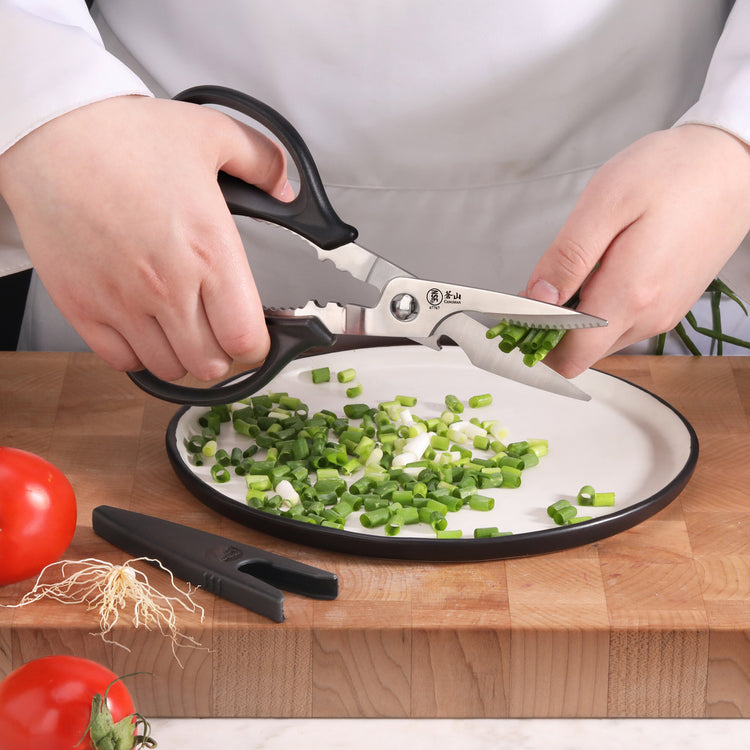 Hands using black-handled kitchen shears to cut green onions over a white plate on a wooden cutting board.