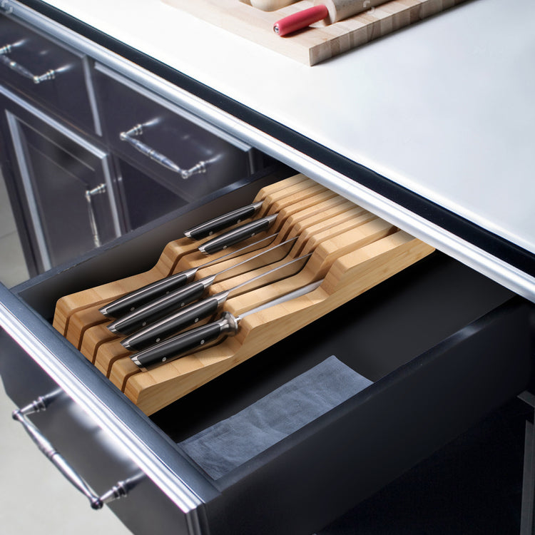 Seven knives with black handles neatly arranged in a bamboo in-drawer knife holder inside a kitchen drawer.