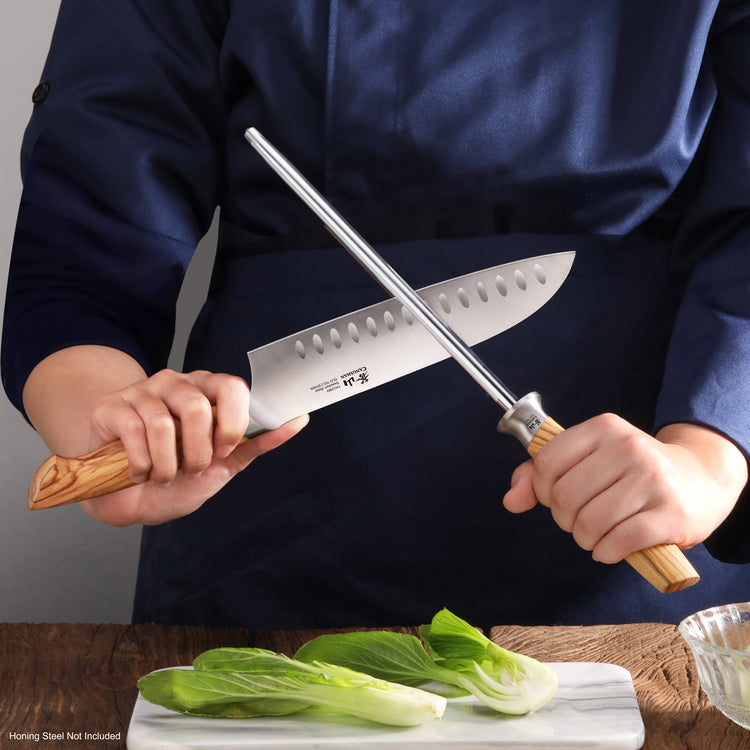 Person sharpening a 7-inch Santoku knife with a wooden handle using a honing steel over a cutting board with green vegetables