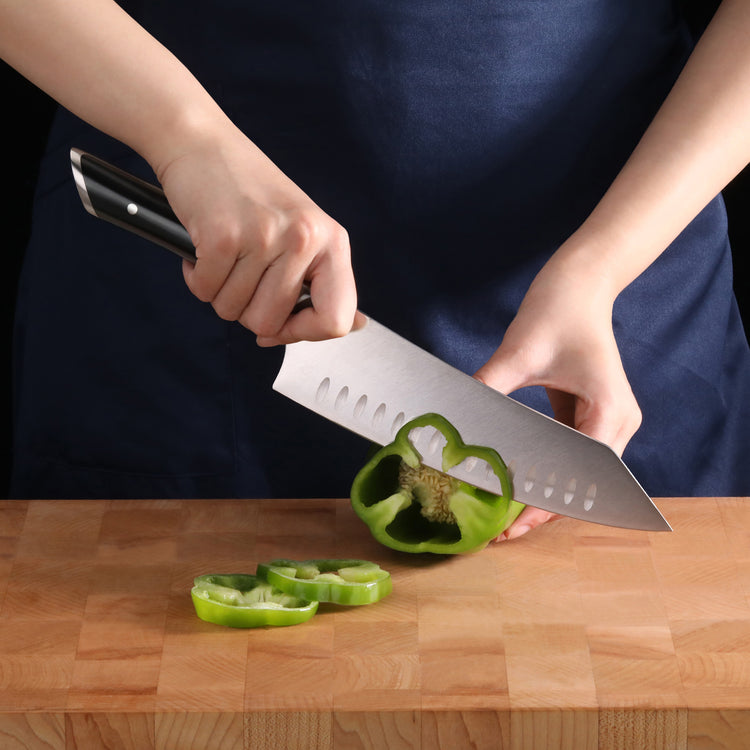 Hand holding a knife slicing a green bell pepper on a wooden cutting board.