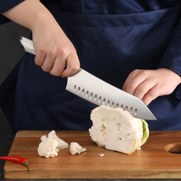 Person slicing a cauliflower head on a wooden cutting board with a large white-handled knife.