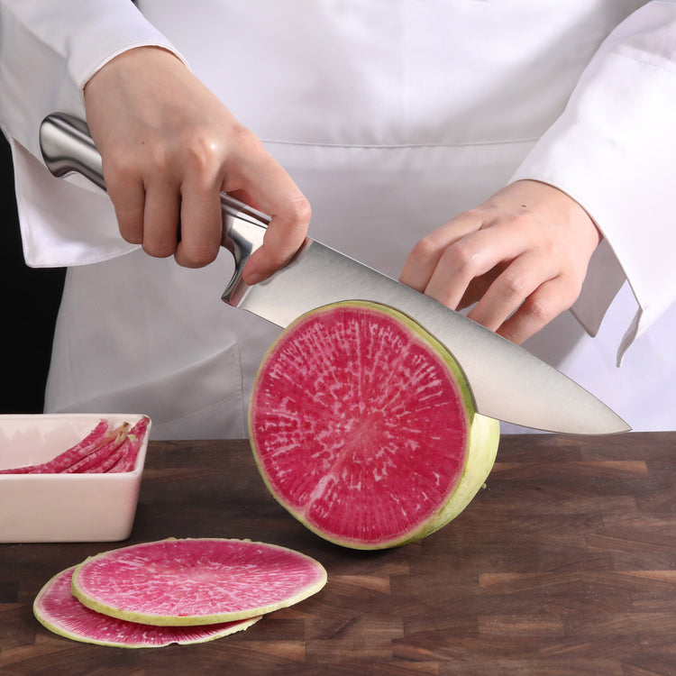 Hands slicing a round pink radish with a large stainless steel chef knife on a wooden cutting board.