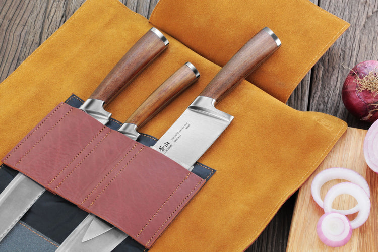 Four knives with wooden handles in a brown leather roll-up case on a wooden surface with sliced onions nearby.