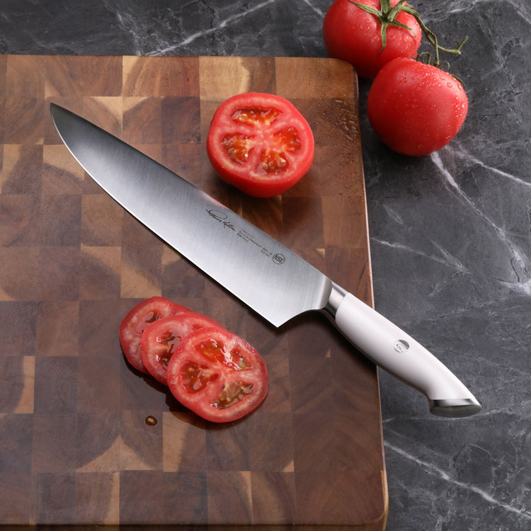 White-handled chef knife on wooden cutting board with sliced and whole tomatoes on dark marble surface.