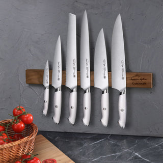 
                    
                      Seven stainless steel knives with white handles mounted on a walnut magnetic knife bar against a gray wall.
                    
                  