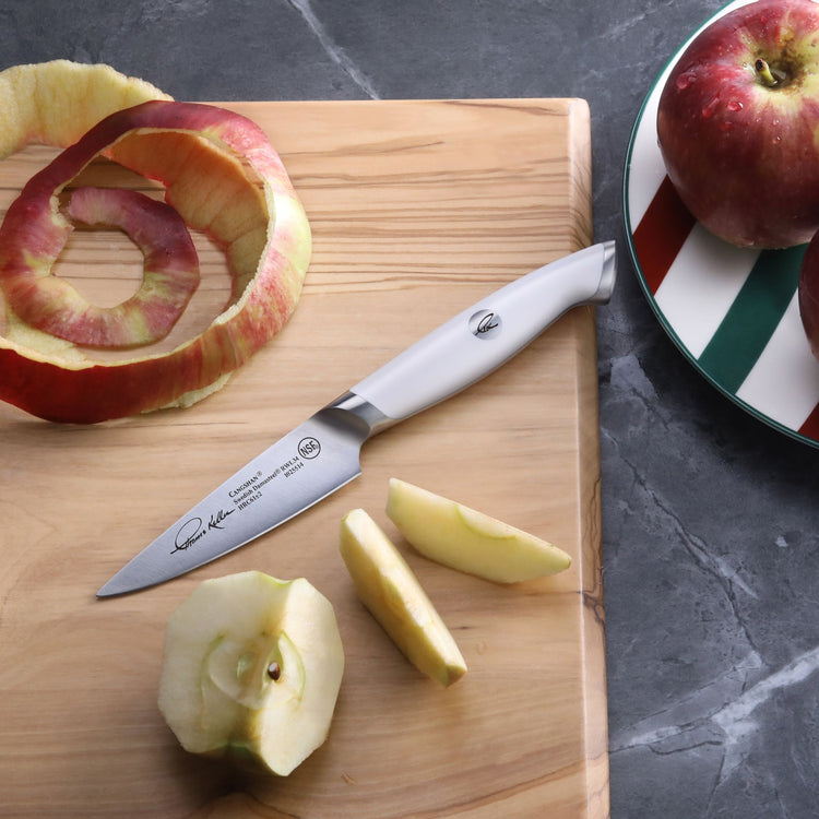 Paring knife with white handle on wooden board next to peeled and sliced apple pieces and apple peel spiral.