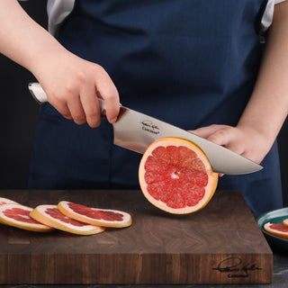 
                    
                      Hand holding a chef's knife cutting a grapefruit on a wooden cutting board with grapefruit slices nearby.
                    
                  